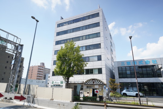 介護施設の建物外観。白い6階建てのモダンな建物で周辺には駐車場と街路樹がある。